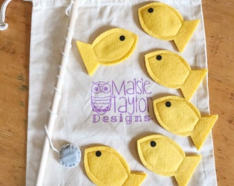 May include: A set of six yellow felt fish with black eyes, a wooden fishing rod, and a gray felt circle. The fish and rod are in a white canvas bag with the text "Maisie Taylor Designs" printed on it.