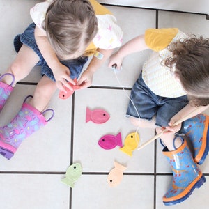 May include: Two children play a fishing game with felt fish on a tiled floor. The fish are pink, yellow, orange, and green. The children are wearing rain boots with colorful patterns.