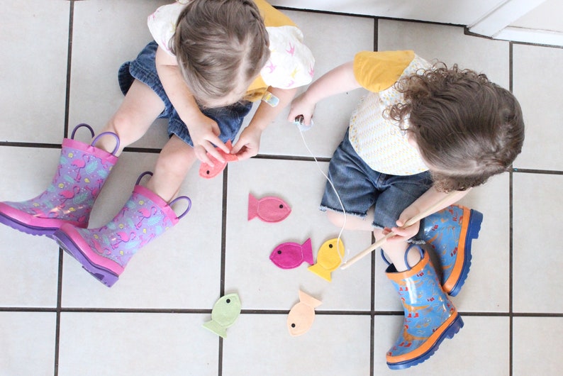 May include: Two children play a fishing game with felt fish on a tiled floor. The fish are pink, yellow, orange, and green. The children are wearing rain boots with colorful patterns.