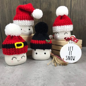 May include: Five white ceramic mugs with smiling faces, each wearing a different knitted Christmas hat. The hats are red, black, and white, some with pom-poms. A white ornament with "LET IT SNOW" is also present.