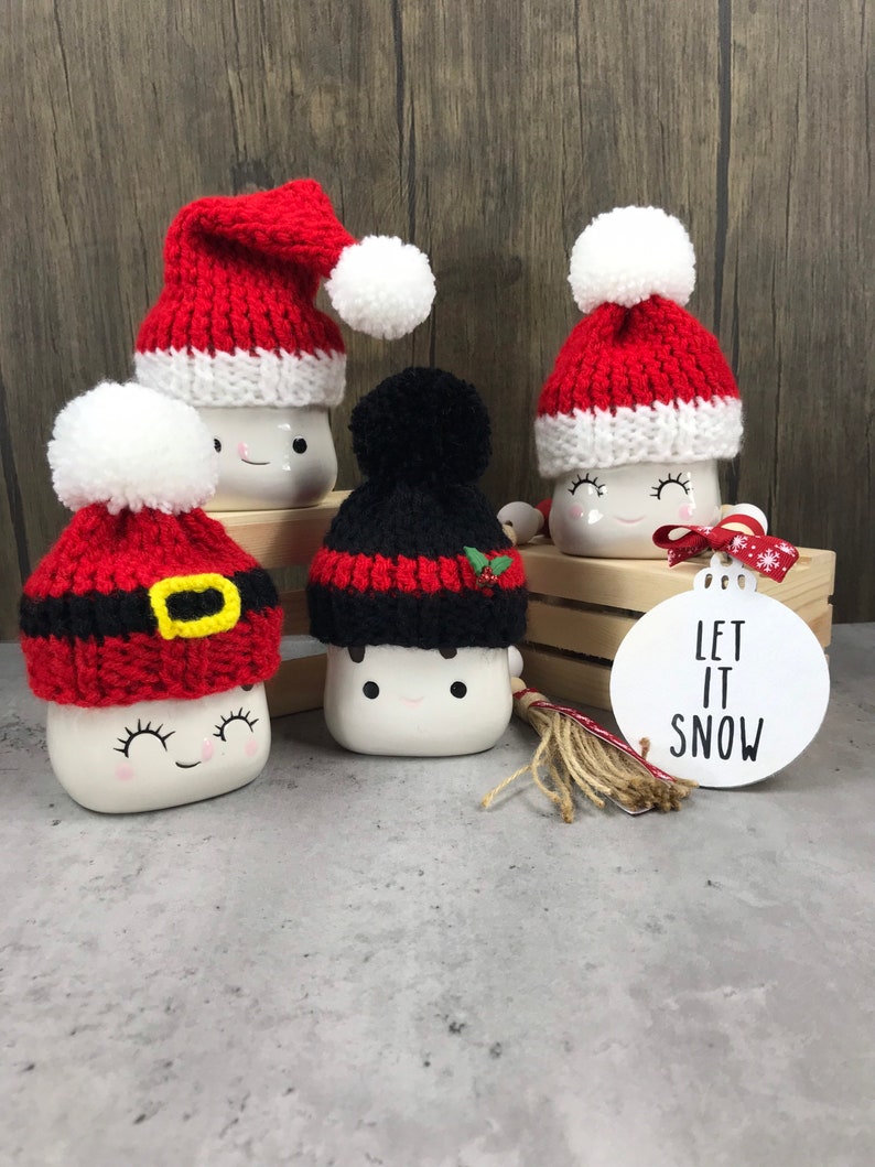 May include: Five white ceramic mugs with smiling faces, each wearing a different knitted Christmas hat. The hats are red, black, and white, some with pom-poms. A white ornament with "LET IT SNOW" is also present.