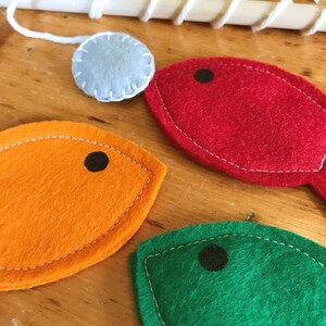 May include: A wooden fishing rod with white string and a light blue felt ball. Three colourful felt fish in red, orange, and green with black dot eyes. The fish are stitched with white thread, creating a playful design.