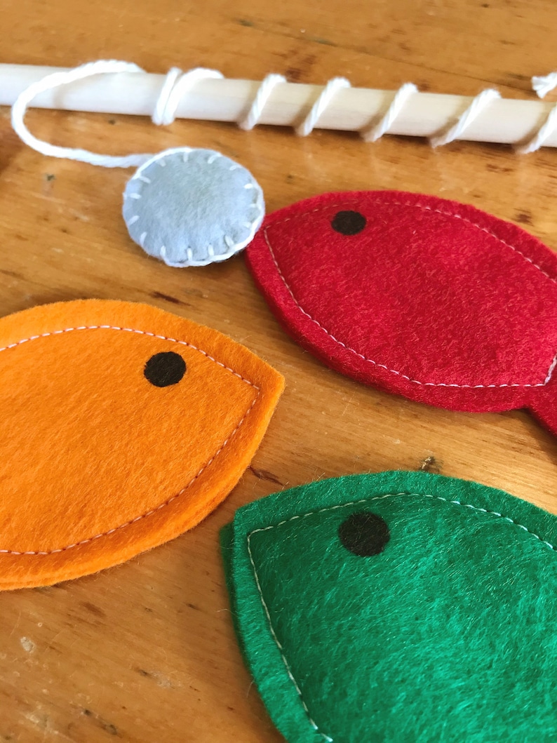 May include: A wooden fishing rod with white string and a light blue felt ball. Three colourful felt fish in red, orange, and green with black dot eyes. The fish are stitched with white thread, creating a playful design.