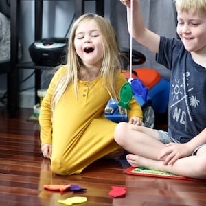 May include: Two children playing a fishing game on a wooden floor. The girl is in a mustard yellow dress, and the boy wears a navy blue t-shirt. There are colourful felt fish scattered on the floor. A blue toy car is in the background.