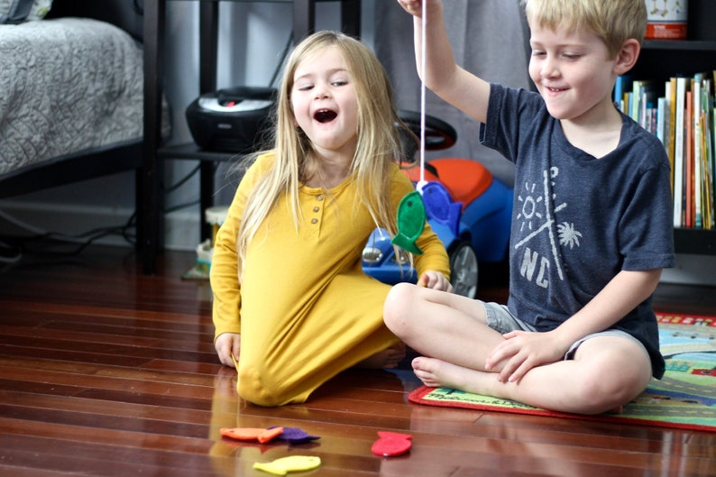 May include: Two children playing a fishing game on a wooden floor. The girl is in a mustard yellow dress, and the boy wears a navy blue t-shirt. There are colourful felt fish scattered on the floor. A blue toy car is in the background.