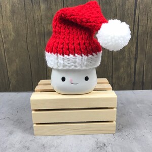 May include: A white ceramic mug with a smiling face, topped with a red and white knitted Santa hat and a white pom-pom. The mug sits on a small wooden crate, with a wooden background.