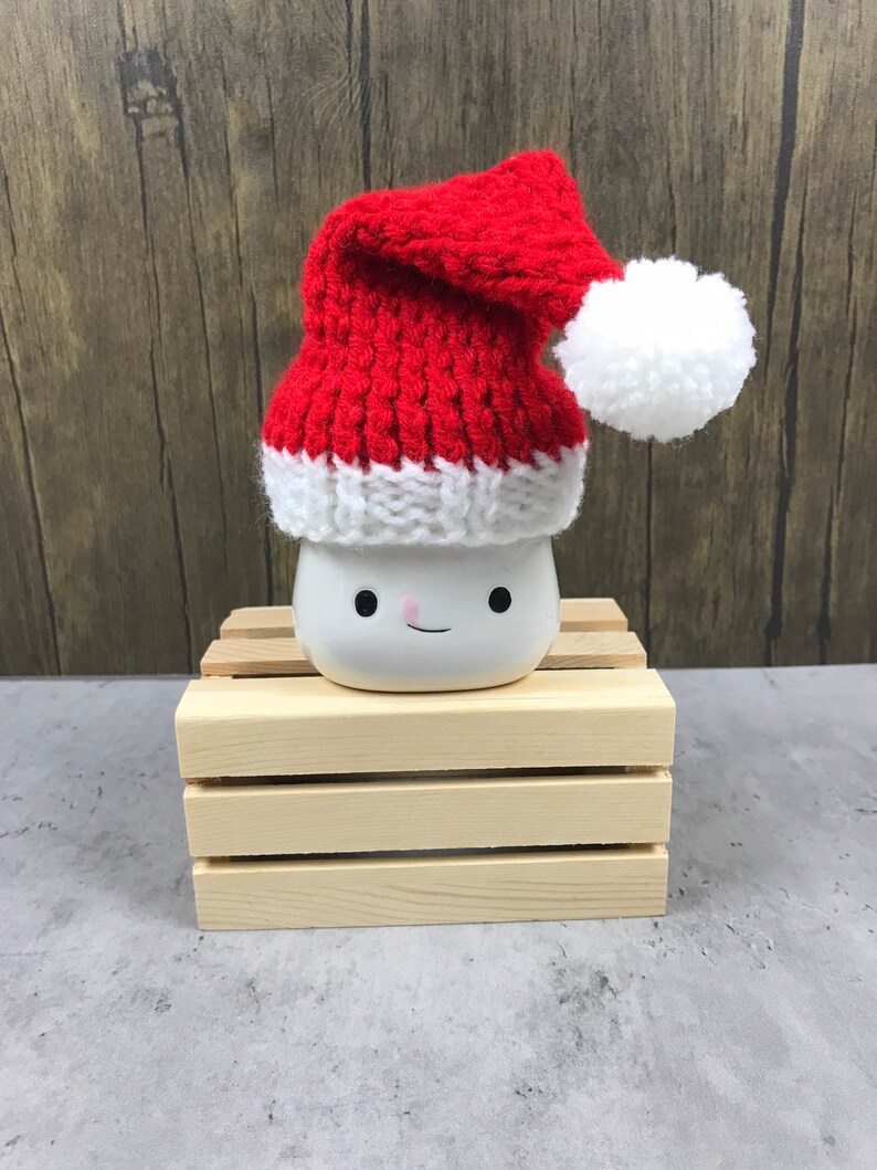 May include: A white ceramic mug with a smiling face, topped with a red and white knitted Santa hat and a white pom-pom. The mug sits on a small wooden crate, with a wooden background.