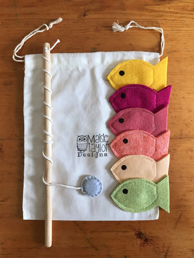 May include: A set of felt fish in various colors, including yellow, pink, red, orange, and green, with black eyes. The fish are attached to a wooden stick with a string and a gray felt circle. The fish are in a white drawstring bag with the text "Maisie Taylor Designs" printed on it.