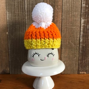 Puede incluir: Una taza de cer&aacute;mica blanca con una cara sonriente que lleva un gorro de punto naranja, amarillo y blanco con forma de ma&iacute;z dulce con un pomp&oacute;n blanco.