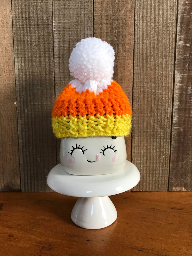 Puede incluir: Una taza de cer&aacute;mica blanca con una cara sonriente que lleva un gorro de punto naranja, amarillo y blanco con forma de ma&iacute;z dulce con un pomp&oacute;n blanco.