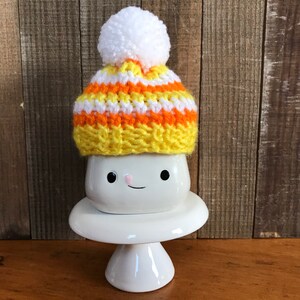 Puede incluir: Una taza de cer&aacute;mica blanca con una cara sonriente, que lleva un gorro de punto de color amarillo, naranja y blanco con un pomp&oacute;n blanco.