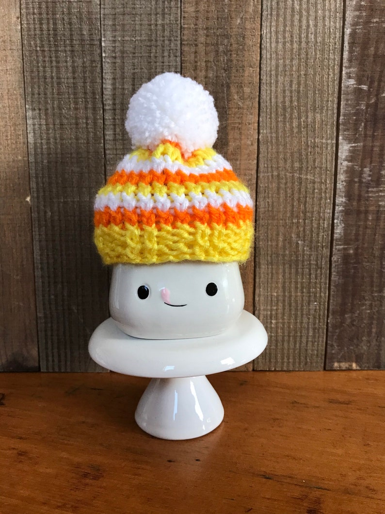 Puede incluir: Una taza de cer&aacute;mica blanca con una cara sonriente, que lleva un gorro de punto de color amarillo, naranja y blanco con un pomp&oacute;n blanco.