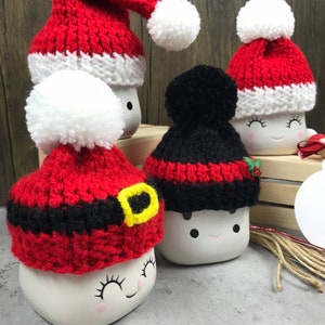 May include: A collection of white ceramic marshmallow mugs, each adorned with a different knitted hat. The hats are red and black with white pom-poms, some with a Santa Claus design. The mugs have painted faces with closed eyes and rosy cheeks.
