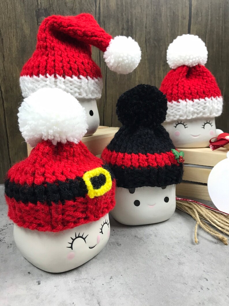 May include: A collection of white ceramic marshmallow mugs, each adorned with a different knitted hat. The hats are red and black with white pom-poms, some with a Santa Claus design. The mugs have painted faces with closed eyes and rosy cheeks.