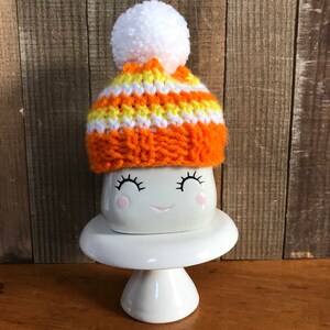 Puede incluir: Una taza de cer&aacute;mica blanca con una cara sonriente y pesta&ntilde;as, que lleva un gorro de punto a rayas naranja y amarillo con un pomp&oacute;n blanco. La taza est&aacute; sentada sobre un pedestal de cer&aacute;mica blanca.