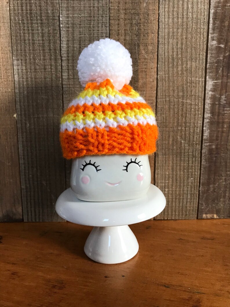 Puede incluir: Una taza de cer&aacute;mica blanca con una cara sonriente y pesta&ntilde;as, que lleva un gorro de punto a rayas naranja y amarillo con un pomp&oacute;n blanco. La taza est&aacute; sentada sobre un pedestal de cer&aacute;mica blanca.