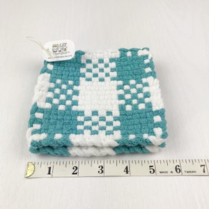 May include: A folded, square, woven washcloth in teal and white. The washcloth features a checkerboard pattern with a white star design. A tag attached to the washcloth reads "Maisie Taylor Designs" and the website address.