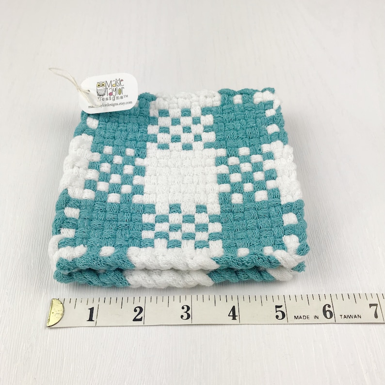 May include: A folded, square, woven washcloth in teal and white. The washcloth features a checkerboard pattern with a white star design. A tag attached to the washcloth reads "Maisie Taylor Designs" and the website address.