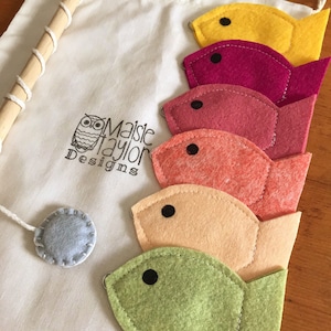 May include: A set of felt fish toys in a variety of colors, including yellow, pink, red, orange, and green. The fish are attached to a wooden dowel rod and come with a gray felt circle. The fish are packaged in a white drawstring bag with the text "Maisie Taylor Designs" printed on it.