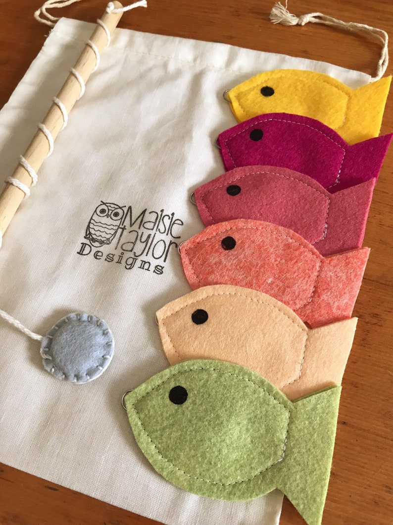 May include: A set of felt fish toys in a variety of colors, including yellow, pink, red, orange, and green. The fish are attached to a wooden dowel rod and come with a gray felt circle. The fish are packaged in a white drawstring bag with the text "Maisie Taylor Designs" printed on it.