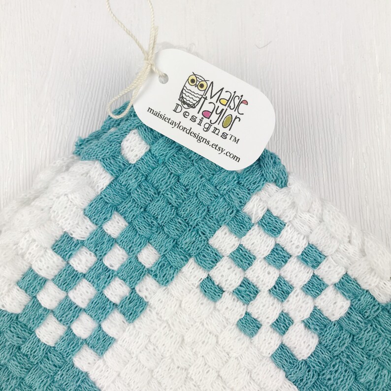 May include: A close-up shot of a woven square in white and teal. The weave creates a geometric pattern. A tag with the Maisie Taylor Designs logo and website address is attached to the top corner of the square.