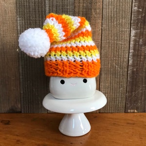 Puede incluir: Una taza de cer&aacute;mica blanca con una cara sonriente, que lleva un gorro de punto de rayas naranja, amarillo y blanco con un pomp&oacute;n blanco. La taza est&aacute; sentada sobre un pedestal de cer&aacute;mica blanca.