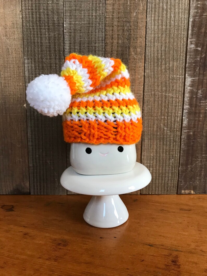 Puede incluir: Una taza de cer&aacute;mica blanca con una cara sonriente, que lleva un gorro de punto de rayas naranja, amarillo y blanco con un pomp&oacute;n blanco. La taza est&aacute; sentada sobre un pedestal de cer&aacute;mica blanca.