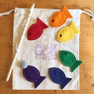 May include: A children's fishing game set. It includes a canvas bag, a wooden fishing rod, and six felt fish in red, orange, yellow, green, blue, and purple. The bag has the text "Maisie Taylor Designs".