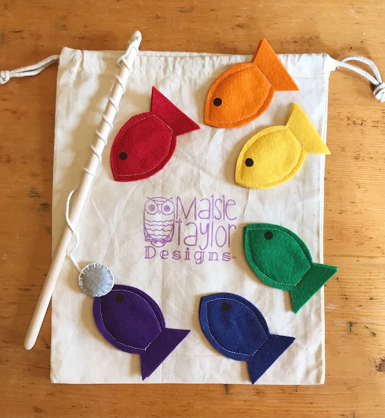 May include: A children's fishing game set. It includes a canvas bag, a wooden fishing rod, and six felt fish in red, orange, yellow, green, blue, and purple. The bag has the text "Maisie Taylor Designs".