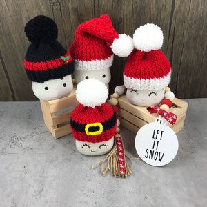 May include: Four marshmallow-shaped Christmas ornaments with knitted hats. One black hat with red stripes and a holly accent, and three red and white Santa hats. A white ornament with "LET IT SNOW" text is also present.