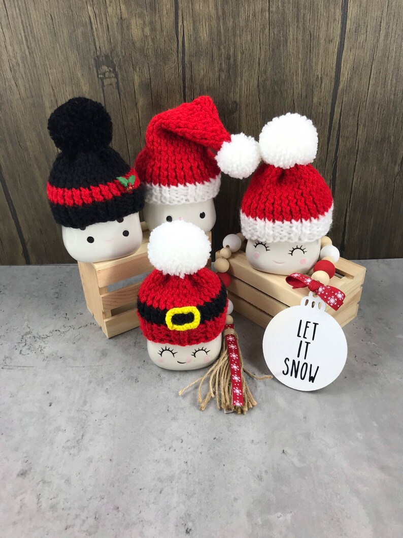 May include: Four marshmallow-shaped Christmas ornaments with knitted hats. One black hat with red stripes and a holly accent, and three red and white Santa hats. A white ornament with "LET IT SNOW" text is also present.
