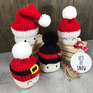 May include: Four ceramic mugs with smiling faces, each adorned with a knitted hat. Three mugs wear red and white Santa hats with white pom-poms, while one has a black and red hat. A white ornament with "LET IT SNOW" is also present.
