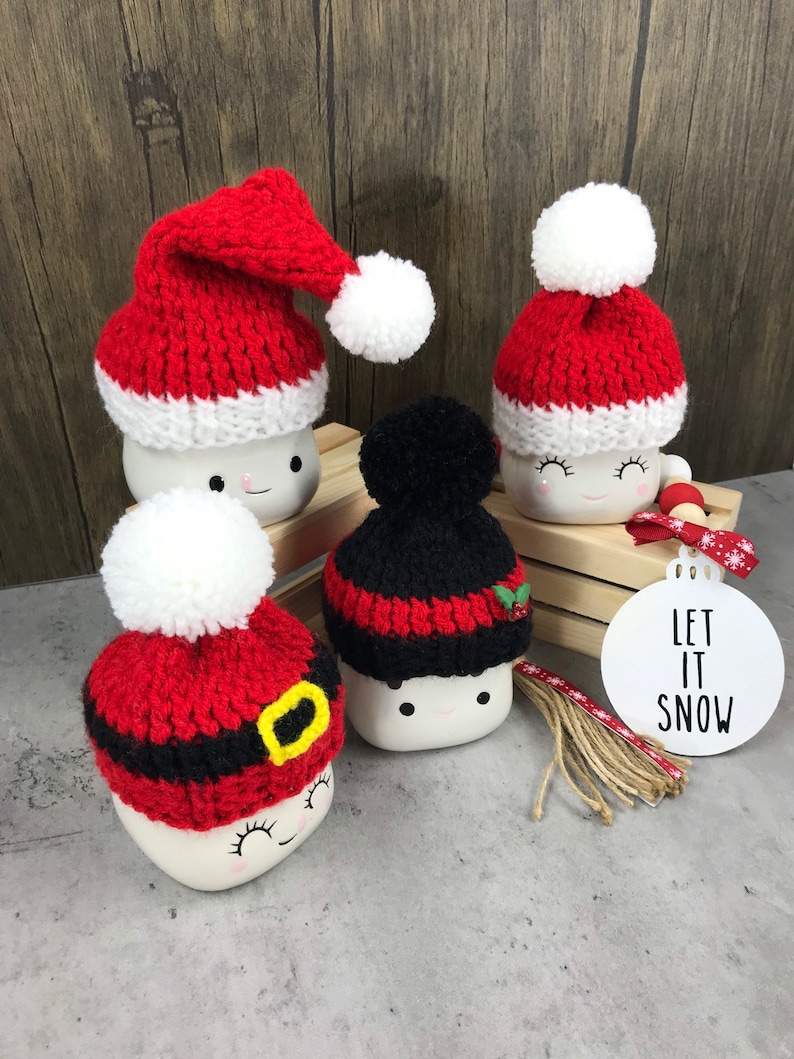 May include: Four ceramic mugs with smiling faces, each adorned with a knitted hat. Three mugs wear red and white Santa hats with white pom-poms, while one has a black and red hat. A white ornament with "LET IT SNOW" is also present.