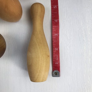 May include: A wooden bowling pin, approximately 5 inches tall, with a smooth, light brown finish. The pin is next to a red measuring tape.