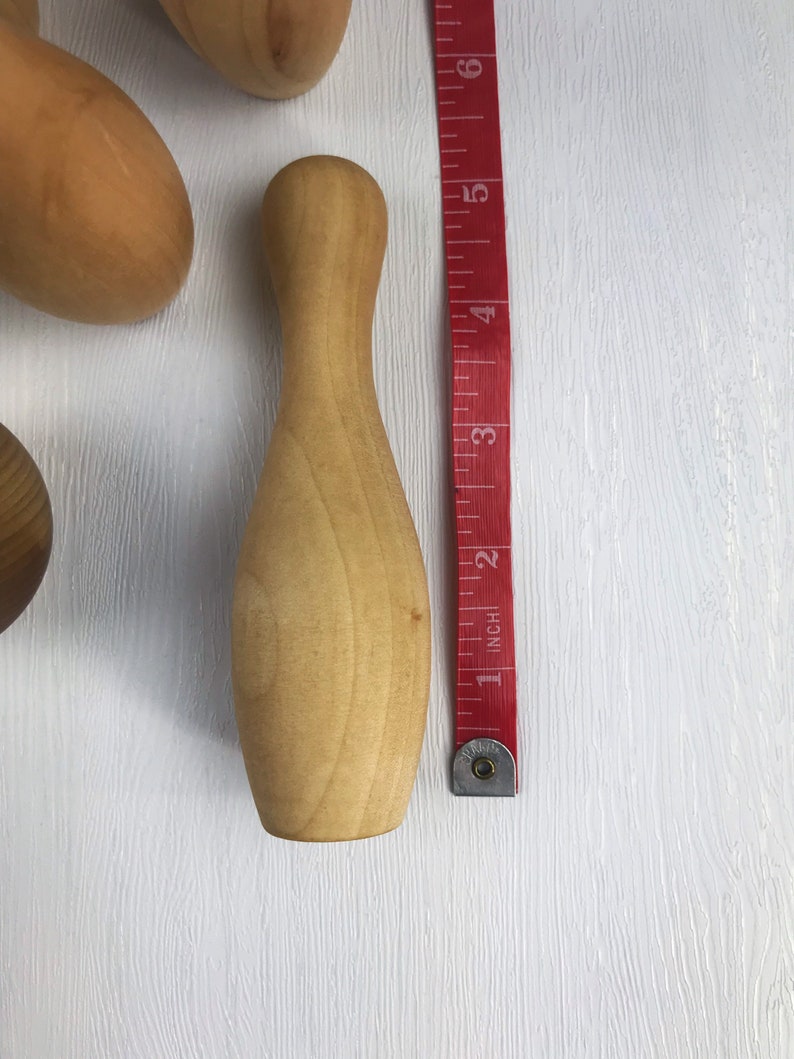 May include: A wooden bowling pin, approximately 5 inches tall, with a smooth, light brown finish. The pin is next to a red measuring tape.
