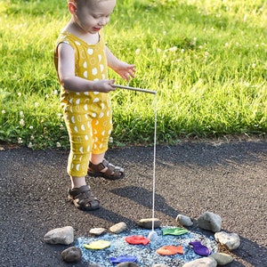 May include: A young child in a yellow and white patterned jumpsuit uses a fishing rod to play a game with colorful fish drawn on the pavement. The child is standing on a black asphalt surface with rocks around the drawn fish.