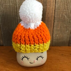 Puede incluir: Un gorro blanco de punto con un dise&ntilde;o de caramelo de ma&iacute;z naranja y amarillo y un pomp&oacute;n blanco en la parte superior. El gorro est&aacute; sentado en una taza blanca con una cara sonriente.