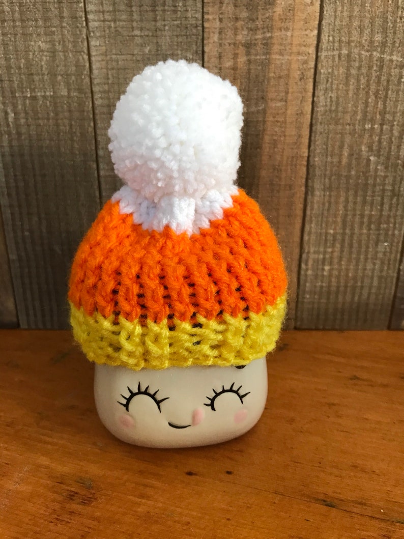 Puede incluir: Un gorro blanco de punto con un dise&ntilde;o de caramelo de ma&iacute;z naranja y amarillo y un pomp&oacute;n blanco en la parte superior. El gorro est&aacute; sentado en una taza blanca con una cara sonriente.