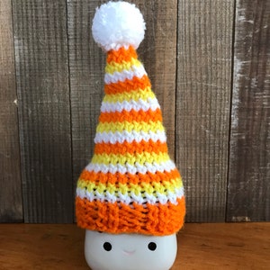 Puede incluir: Una taza de cer&aacute;mica blanca con una cara sonriente que lleva un gorro de punto a rayas naranja, amarillo y blanco con un pomp&oacute;n blanco.