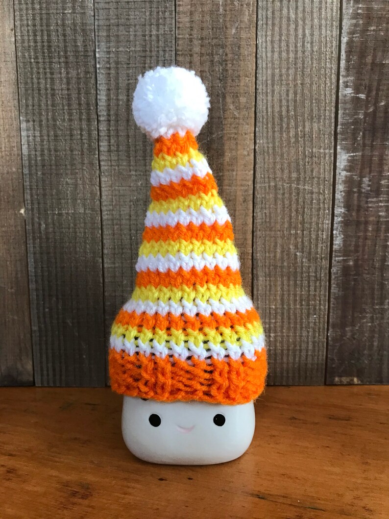 Puede incluir: Una taza de cer&aacute;mica blanca con una cara sonriente que lleva un gorro de punto a rayas naranja, amarillo y blanco con un pomp&oacute;n blanco.