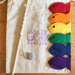 May include: A canvas bag with a drawstring, a wooden fishing rod, and a felt fish game. The bag has a purple owl logo and the text "Maisie Taylor Designs". Six colourful felt fish in red, orange, yellow, green, blue, and purple are arranged on the bag.