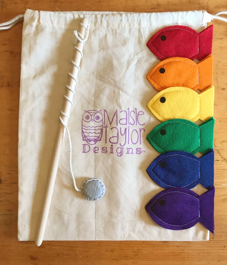 May include: A canvas bag with a drawstring, a wooden fishing rod, and a felt fish game. The bag has a purple owl logo and the text "Maisie Taylor Designs". Six colourful felt fish in red, orange, yellow, green, blue, and purple are arranged on the bag.