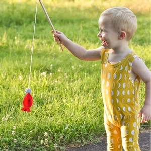 May include: A young child wearing a yellow jumpsuit with a white crescent moon pattern is playing with a red fishing toy. The child is holding a wooden stick with a string attached to a red fish.