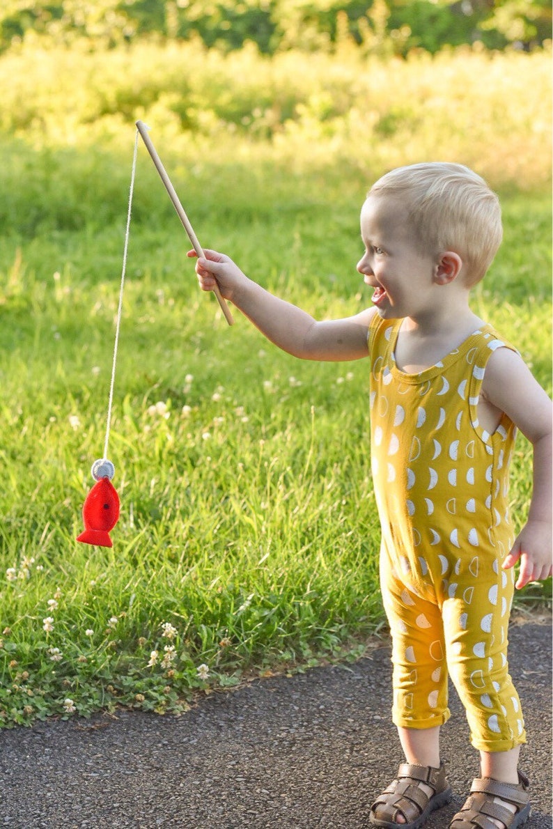 May include: A young child wearing a yellow jumpsuit with a white crescent moon pattern is playing with a red fishing toy. The child is holding a wooden stick with a string attached to a red fish.