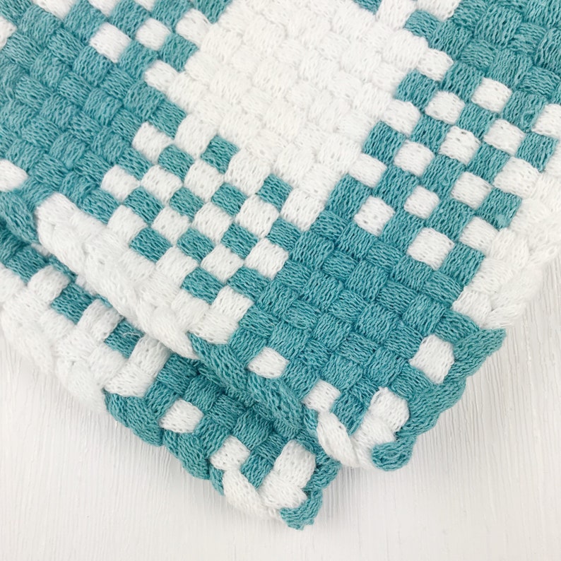 May include: Close-up of two woven potholders. The potholders feature a checkerboard pattern of white and teal squares. The weave is tight, creating a textured surface. The edges are neatly finished, and the potholders are folded over.