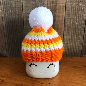 Puede incluir: Una taza de cer&aacute;mica blanca con una cara sonriente que lleva un gorro de punto de rayas naranja, amarillo y blanco con un pomp&oacute;n blanco.