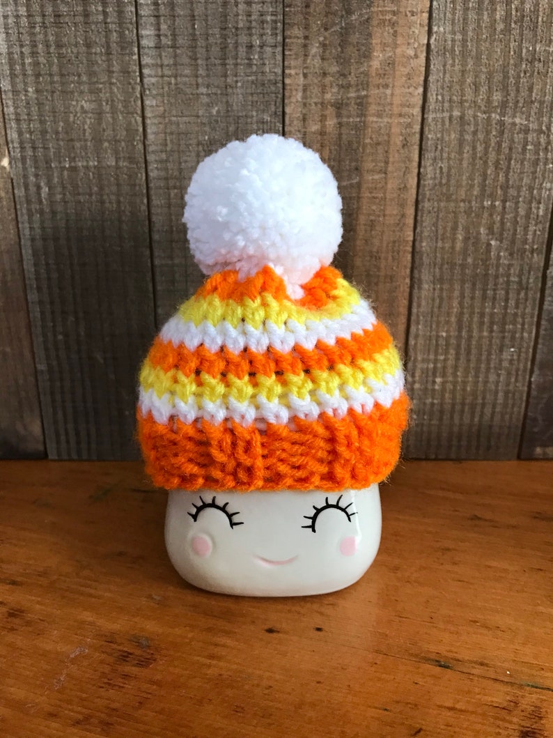 Puede incluir: Una taza de cer&aacute;mica blanca con una cara sonriente que lleva un gorro de punto de rayas naranja, amarillo y blanco con un pomp&oacute;n blanco.