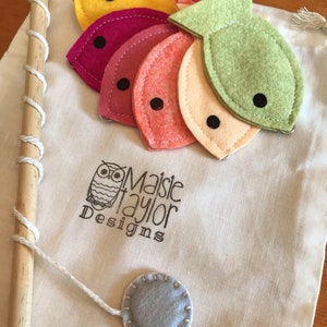May include: A felt fishing game with a wooden pole, string, and five felt fish in different colors: yellow, pink, red, orange, and green. The fish have black eyes and are attached to the string with a small metal ring. The game is packaged in a white canvas bag with the text "Maisie Taylor Designs" printed on it.