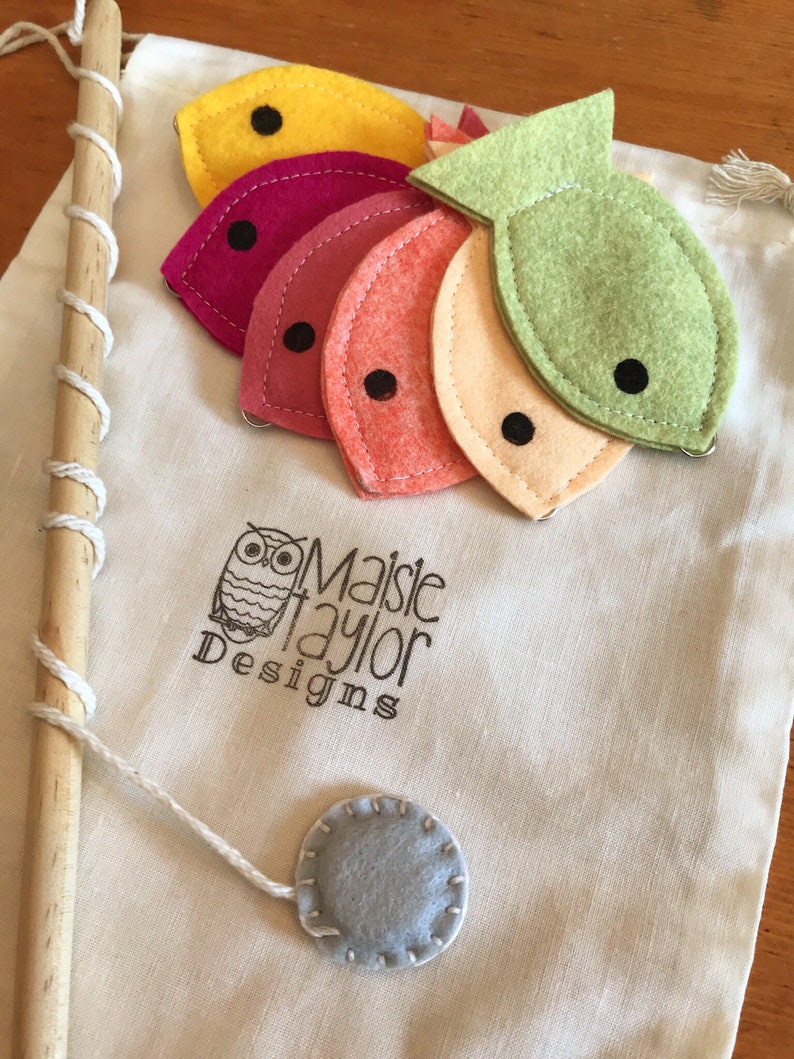 May include: A felt fishing game with a wooden pole, string, and five felt fish in different colors: yellow, pink, red, orange, and green. The fish have black eyes and are attached to the string with a small metal ring. The game is packaged in a white canvas bag with the text "Maisie Taylor Designs" printed on it.