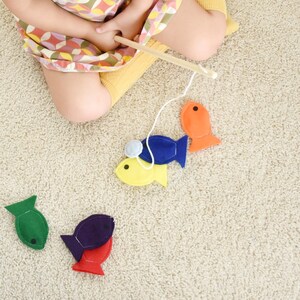 May include: A child's fishing game featuring felt fish in various colours: green, purple, red, yellow, blue, and orange. A wooden fishing rod with a white string is held above the fish. The game is set on a beige carpet.